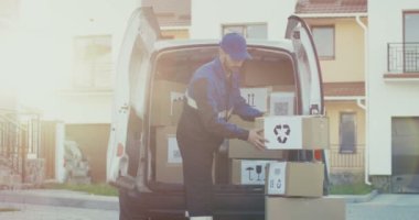 Beyaz erkek kurye mavi kostümlü ve kepli güneşli bir günde beyaz minibüsten posta kutularını alıyor. Açık hava..