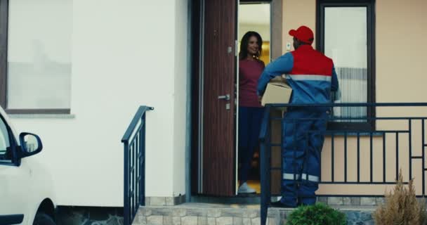 African American mailman in the uniform ringing in the doorbell, Caucasian woman opening a door and getting a parcel. Outside. — Stock Video