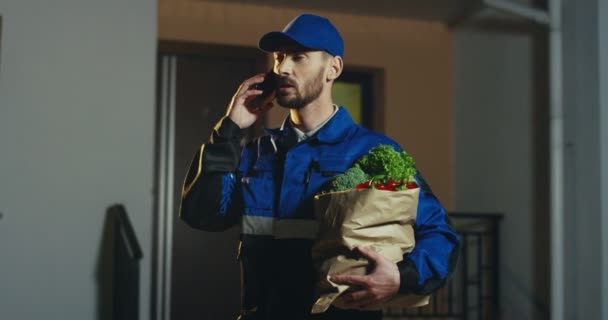 Kavkazský atraktivní superamrket poslíček v modrém kostýmu a čepici s čerstvou zeleninou v papírovém obalu, který večer mluví na mobilním telefonu. Vně. — Stock video