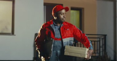 Kırmızı üniformalı Afro-Amerikalı genç teslimatçının portre resmi ve yan tarafa bakan bir paket ile bir şapka ve sonra gece kameraya gülümsüyor. Açık havada..