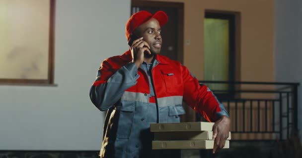 Afroameričtí pracovníci na pizzu dodávky v červené uniformě a čepici stojí v domě v noci s krabicemi od pizzy a mluví na mobilním telefonu v. Outdoor. — Stock video