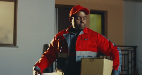 Портрет молодого и веселого афроамериканского почтальона в красном костюме и кепка с коробкой, смотрящей в сторону, а вечером поворачивающей голову к камере. Снаружи. — стоковое видео