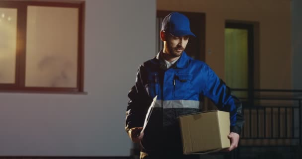 Retrato del repartidor caucásico guapo con el uniforme azul y una gorra con un paquete mirando y luego sonriendo a la cámara por la noche. Al aire libre. — Vídeo de stock