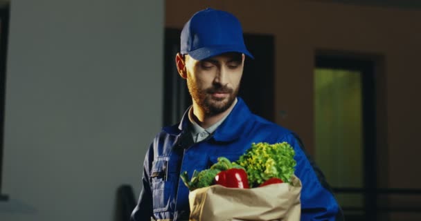 Primer plano del atractivo trabajador masculino caucásico de la entrega del supermercado posando y sonriendo a la cámara con un paquete de cartón lleno de verduras frescas. Afuera. Retrato por la noche. — Vídeo de stock
