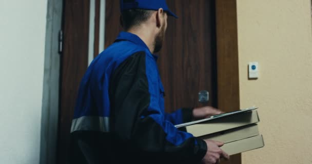 Zadní strana kavkazského poslíčka pizzy v modré uniformě zvoní na zvonek, krásná žena otevírá dveře, podepisuje a přijímá objednávku. Vnitřní. — Stock video