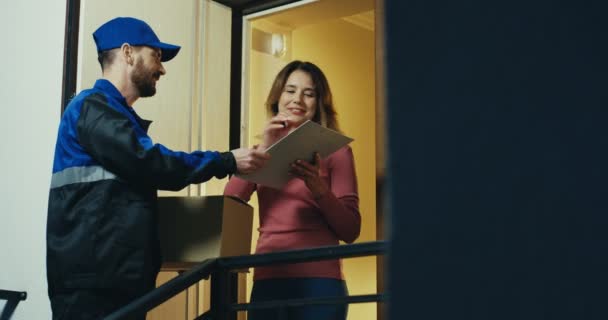 Postino caucasico in uniforme blu che suona al campanello, bella donna che apre una porta, firma e prende la sua casella di posta. Interni. — Video Stock