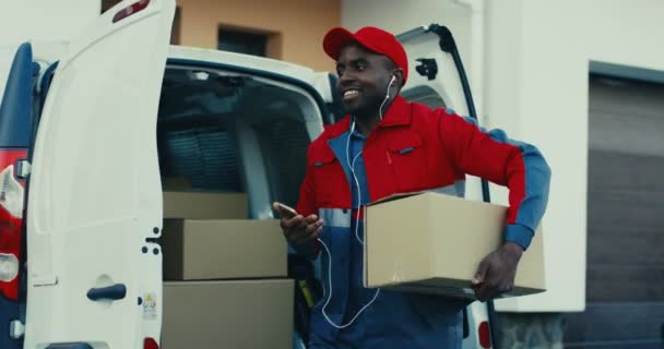 Веселый молодой афроамериканец в красной форме достает коробку из фургона и идет к дому, слушая музыку в наушниках и улыбаясь. На открытом воздухе. — стоковое видео