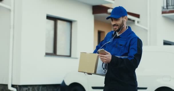 Alegre y atractivo repartidor caucásico en el uniforme azul caminando con un paquete en las manos y bailando como escuchar la música un auriculares. Exterior. — Vídeos de Stock