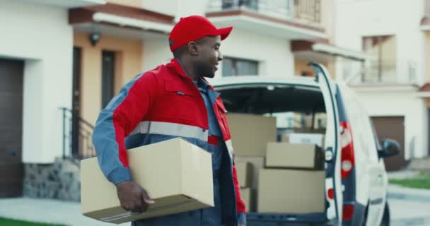 Портрет молодого улыбчивого афроамериканского почтальона с коробкой, позирующей перед камерой с коробкой в фургоне. На открытом воздухе. — стоковое видео