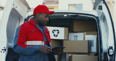 Kırmızı üniformalı ve elinde tabletle posta kutularını kontrol eden Afro-Amerikan nakliye şirketinde çalışan erkek işçiyi kapatın. Dışarıda..