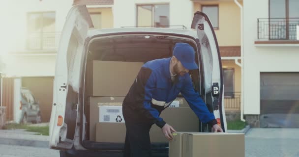 Knappe blanke mannelijke werknemer van de rederij in het rode kostuum en pet die kartonnen pakketten uit het busje haalt. Buiten. — Stockvideo