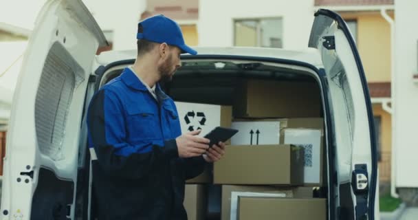 Белый симпатичный почтальон в синем костюме и кепке проверяет почтовые ящики с планшетом в руках. На улице. Камера приближается.. — стоковое видео