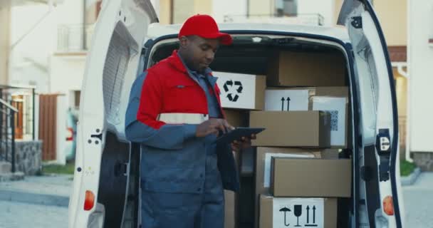 Афроамериканец в красном костюме и кепке проверяет почтовые ящики с планшетным компьютером в руках. На улице. Камера приближается.. — стоковое видео