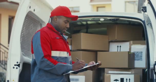 Retrato del joven trabajador afroamericano de la naviera parado en la furgoneta con cajas y escribiendo el documento, luego sonriendo a la cámara. Al aire libre. — Vídeo de stock