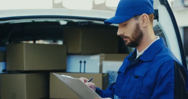 Primer plano del atractivo trabajador masculino caucásico escribiendo un documento en la furgoneta llena de paquetes de cartón. Afuera.. — Vídeo de stock