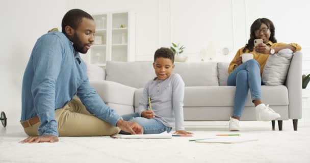 Padre joven afroamericano jugando con su hijo pequeño con lápices en el suelo en la sala de estar. Concepto de dibujo. Madre usando el teléfono inteligente y sentado en el sofá en el fondo Papá y el niño pasar tiempo juntos — Vídeo de stock