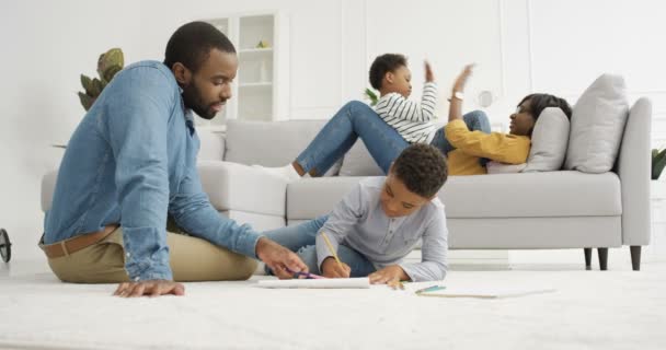 Padre afroamericano con hijo pequeño para colorear cuadro con lápices en el suelo en la sala de estar. Madre jugando juego de manos con la hija en el sofá en el fondo. Padres e hijos pasando tiempo juntos. — Vídeos de Stock