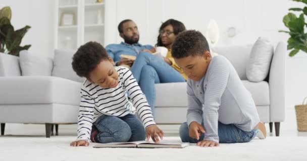 Afro-américains heureux enfants assis sur le sol et feuilletant des pages de livre. Petite sœur et petit frère jouant à la maison. Deux parents assis sur le canapé et parlant en arrière-plan. Repos de père et mère. — Video