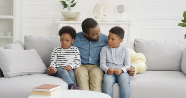 Ung afroamerikansk stilig far sitter på soffan med två små barn och pratar. Pappa kramar små barn på sfa och berättar sagor eller predikar. Hemma. Man med son och dotter. — Stockvideo