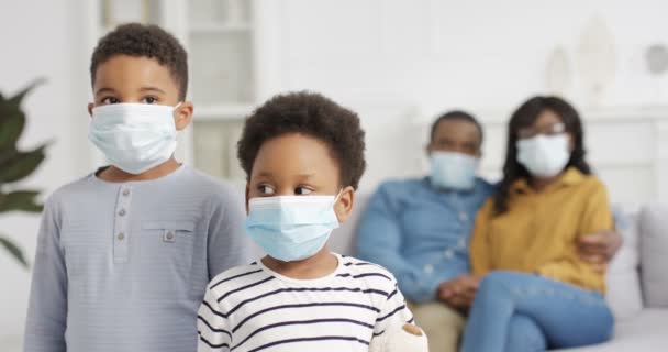 Portrét malých afroamerických dětí v lékařských maskách, jak se doma dívají do kamery. Rodiče sedí na pohovce na rozmazaném pozadí. Malá roztomilá sestra a bratr v karanténě. Koncept koronaviru. — Stock video