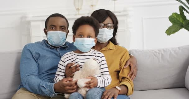 African American ung familj med söt liten unge i medicinska masker hemma på lockdown. Lycklig far, mor och liten dotter sitter på soffan i kramar i vardagsrummet. Begreppet coronavirus. — Stockvideo