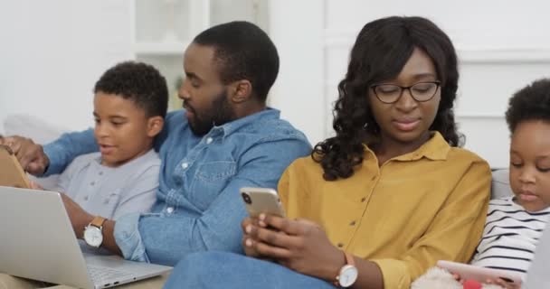 Afroameričtí rodiče s dětmi na gauči pomocí pomůcek. Mladý muž a žena se synem a dcerou tráví čas spolu s pomůckami Otec a syn s notebookem Matka a dcera s chytrými telefony — Stock video