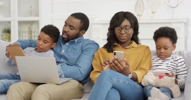 在平板电脑上和儿子玩笔记本电脑的非裔美国父亲。妈妈和女儿用智能手机。一家人在家里沙发上呆着。有孩子和小玩艺儿的父母. — 图库视频影像