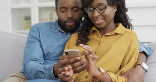 Joven pareja alegre afroamericana sentada en el sofá y viendo algo en la pantalla del teléfono inteligente. En casa. Hombre guapo y mujer hermosa usando el teléfono móvil, tocando y desplazándose. De interior. — Vídeos de Stock