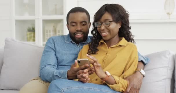 Afro-Amerikaanse jonge knappe man glimlachend en knuffelend mooie vrouw. Gelukkig stel zit op de bank en kijkt thuis iets op de mobiele telefoon. Vriendin en vriendje met smartphone. — Stockvideo