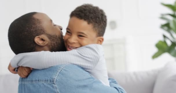 Feliz hombre afroamericano alegre abrazando a su pequeño hijo lindo y riéndose en casa en el sofá. Padres alegres con niños pequeños en el salón. Padre joven sonriendo y abrazando al niño en el interior. Generaciones — Vídeo de stock