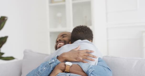 Feliz hombre afroamericano alegre abrazando a su pequeño hijo lindo y riéndose en casa en el sofá. Padres alegres con niños pequeños en el salón. Padre joven sonriendo y abrazando al niño en el interior. Parte trasera. — Vídeos de Stock