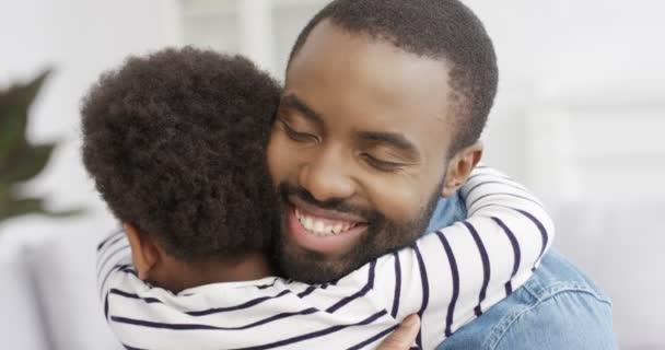 Feliz homem afro-americano alegre abraçando sua pequena filha fofa e rindo de casa. Pais alegres com a criança na sala de estar. Jovem pai sorrindo e abraçando criança interior. Traseira da menina. — Vídeo de Stock