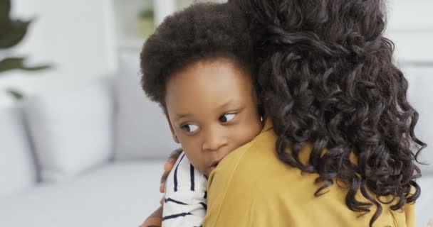 Πίσω από χαρούμενη Αφρο-Αμερικανίδα που αγκαλιάζει την κορούλα της στον καναπέ. Γυναίκα με σγουρά μαλλιά και μικρό παιδί στο σαλόνι. Πλάτη άποψη για τη μητέρα χαμογελά και αγκαλιάζει το παιδί — Αρχείο Βίντεο