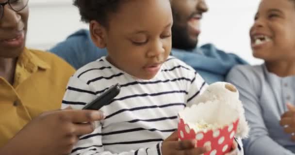 Felice famiglia afroamericana trascorrere il tempo libero insieme a casa. Piccoli bambini carini con i genitori che mangiano popcorn e guardano la TV. Madre e padre si divertono con piccolo figlio e figlia. — Video Stock