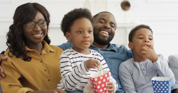 Glada afroamerikanska föräldrar med små barn som sitter på soffan, skrattar, tittar på komedi film och äter popcorn. Söta små barn med mamma och pappa umgås hemma. — Stockvideo