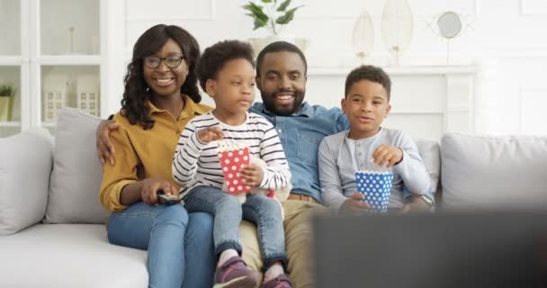 Feliz família afro-americana sentada no sofá na sala de estar e assistindo TV junto com pipocas. Crianças bonitas com mãe alegre sorridente e pai se divertindo juntos em casa. Parentalidade — Vídeo de Stock