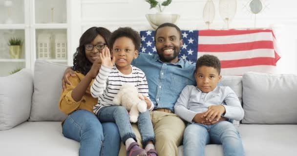 Porträtt av unga glada afroamerikanska föräldrar barn sitter på soffan med USA flagga och ler glatt mot kameran. Glada barn med mor och far på soffa i vardagsrummet. — Stockvideo