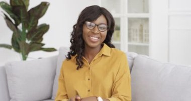 Güzel Afro-Amerikan kadın blogcu oturma odasında kanepede oturmuş kamerayla konuşuyor. Kadın video kaydedicisi bir şey söylüyor ve el sallıyor. Video blogu. Evden video bloğu. Videochat konuşması.