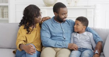 Neşe dolu Afrikalı Amerikalı bir ailenin küçük sevimli bir çocukla portresi. Küçük çocuk oturma odasında ailesiyle birlikte koltukta oturuyor ve kameraya mutlu bir şekilde gülümsüyor. Anne, baba ve oğlu evdeki kanepede..