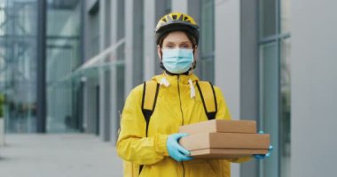 Güzel beyaz kadın teslimatçının portre fotoğrafı. Casque içinde, eldivenler ve tıbbi maske takıyor ve elinde karton kutular var. Kameraya bakan güzel bir kadın kurye..