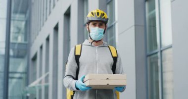 Casque giymiş yakışıklı beyaz bir teslimatçının portre fotoğrafı, eldivenler ve tıbbi maske sokakta duruyor ve elinde karton kutular var. Pizza yiyen erkek kurye kameraya bakıyor..