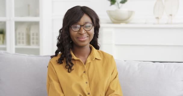 Retrato de una hermosa joven afroamericana con camisa amarilla y gafas sentadas en el sofá y sonriendo a la cámara. Bonita mujer guapa con sonrisa en el sofá en casa. — Vídeo de stock