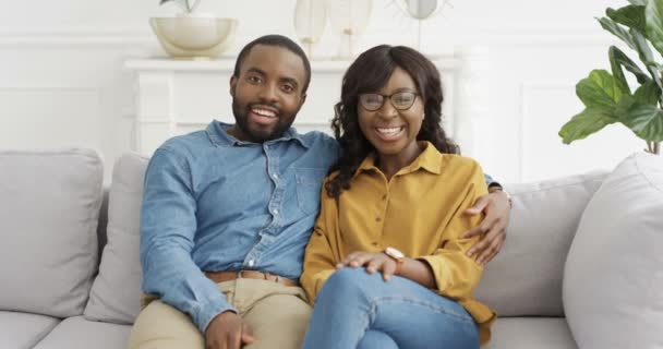 Retrato de una joven pareja afroamericana feliz sentada en abrazos en el sofá y hablando con la cámara como blogueros. Mujer bonita y hombre guapo sonriendo alegremente y diciendo algo. Videochat en casa. — Vídeos de Stock