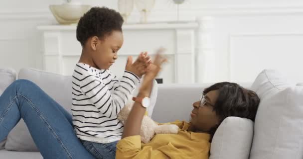 Afro-Amerikalı mutlu anne evdeki kanepede küçük tatlı kızıyla el oyunu oynuyor. Anne ve küçük çocuk oturma odasında kanepede birlikte eğleniyorlar. Annelik ve çocukluk kavramı. — Stok video