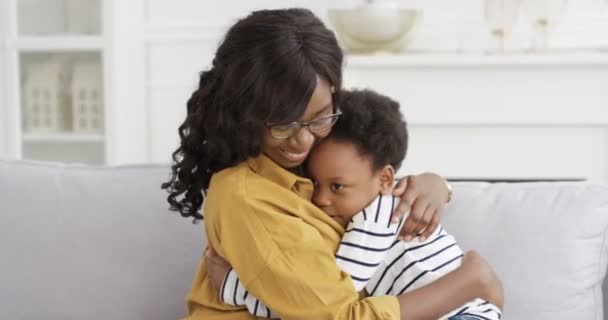 Afro-Amerikaanse jonge mooie vrouw met bril zitten op de bank thuis en knuffelen haar schattige kleine dochter. Klein meisje omarmt moeder op de bank in de woonkamer. Moederliefde. Gelukkige jeugd. — Stockvideo