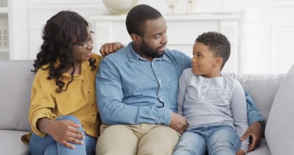 Ritratto di giovane famiglia afroamericana felice con piccolo figlio carino. Ragazzino seduto sul divano in salotto con i genitori e sorridente gioiosamente. Madre e padre con bambino sul divano a casa. — Video Stock