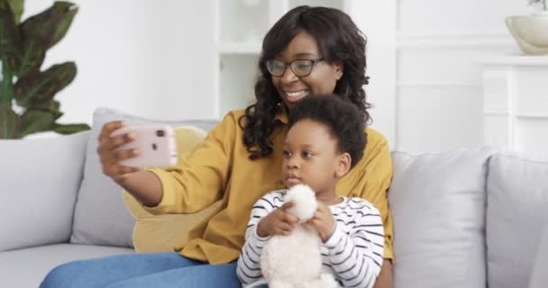 Mooie jonge Afro-Amerikaanse moeder zit op de bank met schattige kleine dochter en het nemen van selfie foto 's met smartphone camera. Thuis. vrouw met klein kind het maken van foto 's selfies met telefoon. — Stockvideo