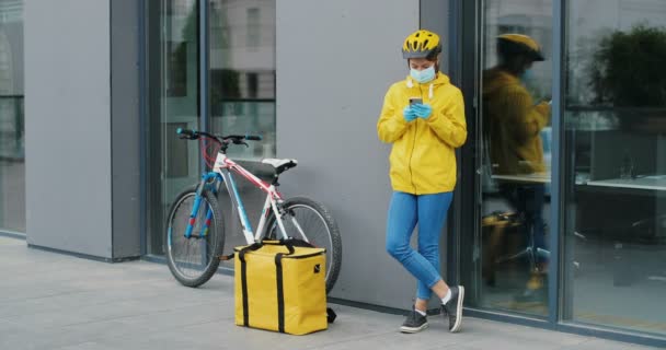 自転車の屋外に立っている医療用マスクの白人女性配達労働者とスマートフォンでタップまたはスクロールします。フードランニング中の男泥棒バッグをつかんで逃げる。女性からの供給を盗む. — ストック動画