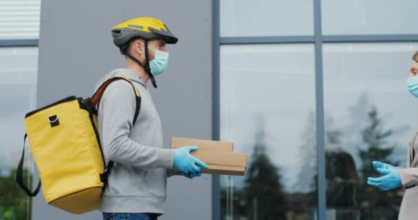 Bělošský mladý doručovatel v casque, rukavice a lékařská maska dávající balíky ženě na ulici. Muž byciclist s taškou přinášející krabice na ženské zákaznice venkovní. — Stock video