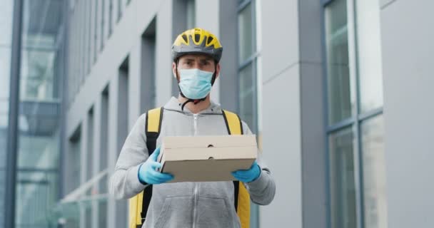 Retrato de repartidor caucásico guapo en casco, guantes y máscara médica de pie en la calle, sosteniendo y entregando cajas de cartón. mensajero masculino dando pizza y mirando a la cámara. — Vídeos de Stock